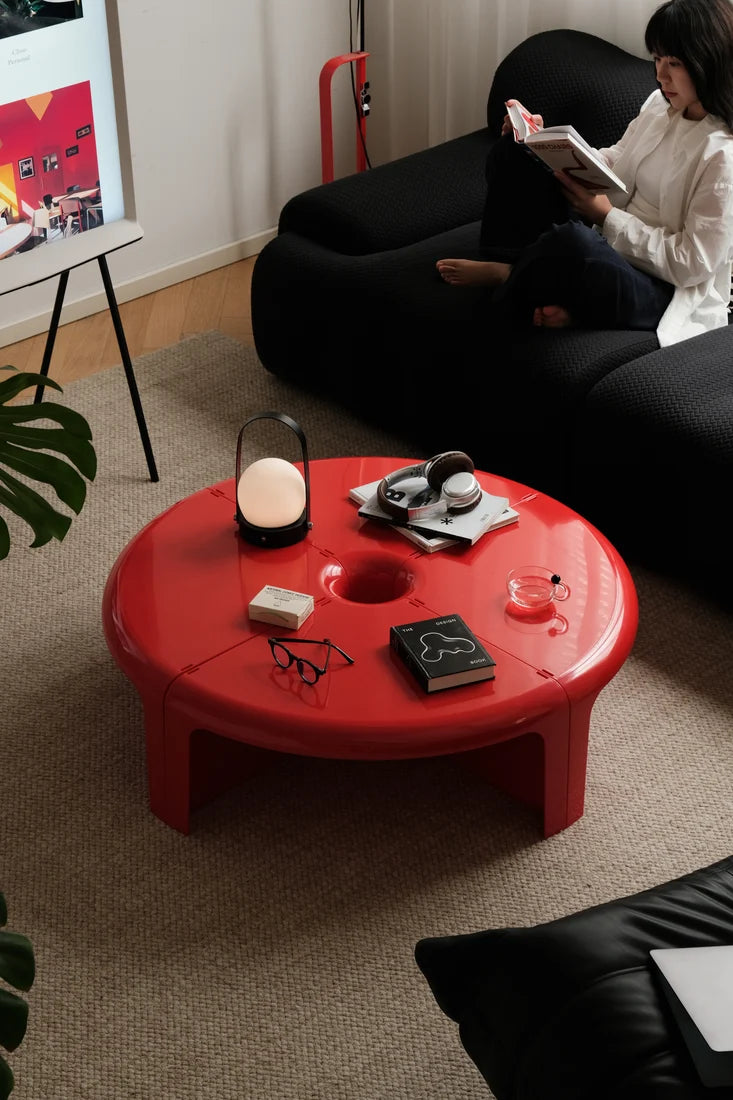 A bright red round low coffee table in a modern living room setting, styled with a glass mug, a small lamp, and other accessories. A woman is seated on a black sofa in the background reading a book.