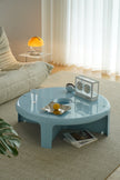 A round light blue coffee table in a modern living room setting, with a person's hand reaching for a cup on the table. Books are placed on the table, including an Alto magazine and a book with a red abstract cover.