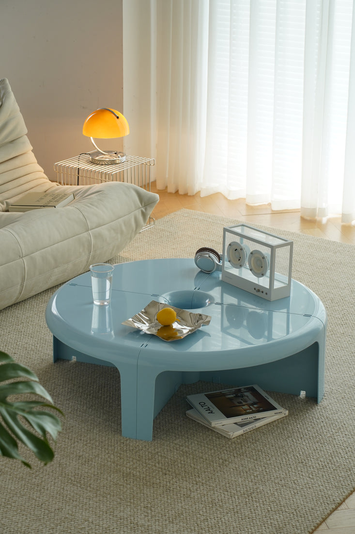 A round light blue coffee table in a modern living room setting, with a person's hand reaching for a cup on the table. Books are placed on the table, including an Alto magazine and a book with a red abstract cover.