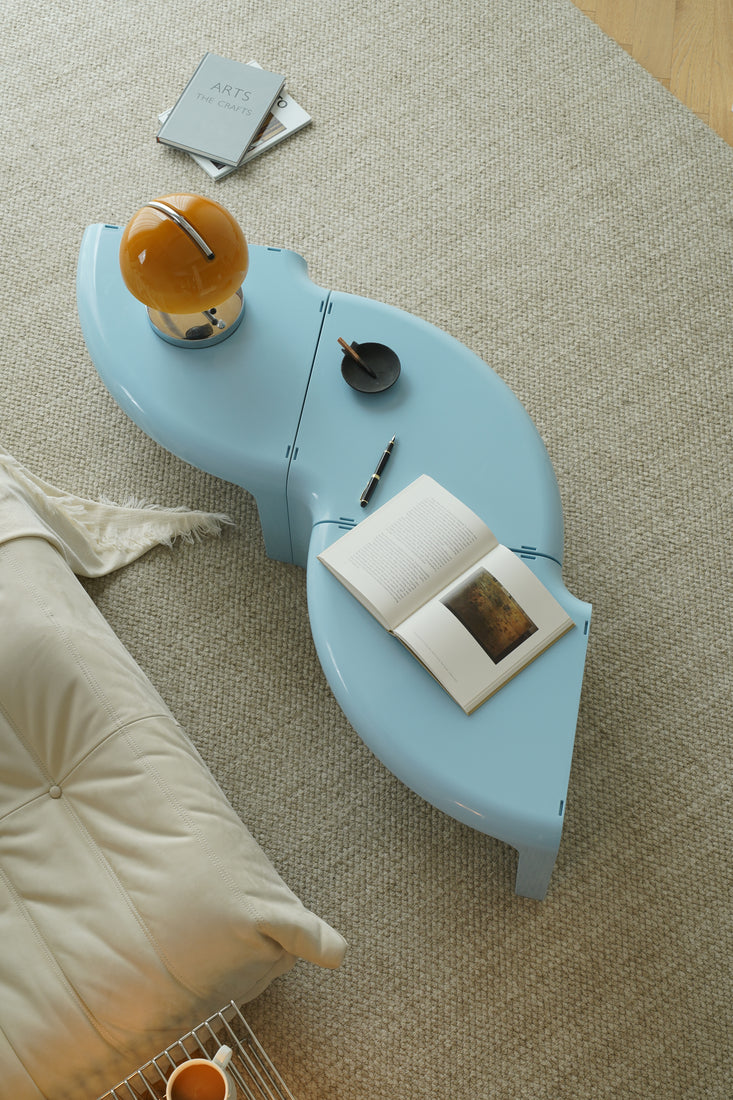 A light blue low coffee table in a modern living room setting, styled with a glass mug, a small lamp, and other accessories. A woman is seated on a black sofa in the background reading a book.