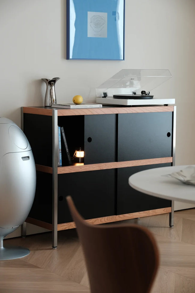 A modern two-tone cabinet with black doors and light wood trim, styled in a minimalist room setting with a turntable on top, a small lamp, and a vase.