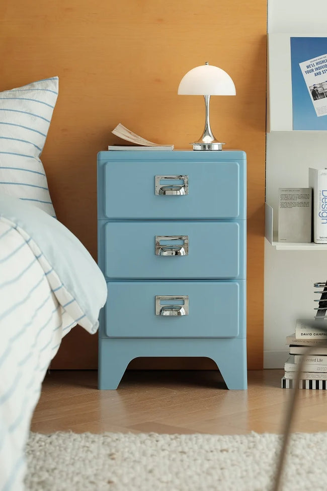 A light blue three-drawer nightstand with silver handles, styled in a bedroom next to a bed with blue-striped bedding, a white table lamp, and a wooden wall background.