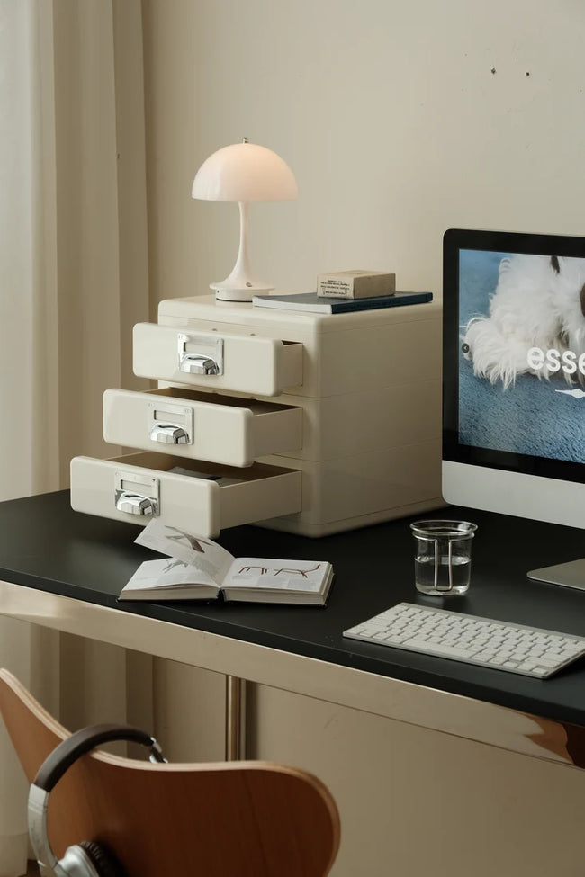 A cream-colored three-drawer desktop organizer with silver handles sits on a black desk next to a computer monitor, keyboard, lamp, and an open notebook in a styled home office setting.
