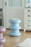 A light blue, tiered barrel-style side table with a glossy finish, styled with a magazine on top in a bright, modern room with light wood floors and white furniture.