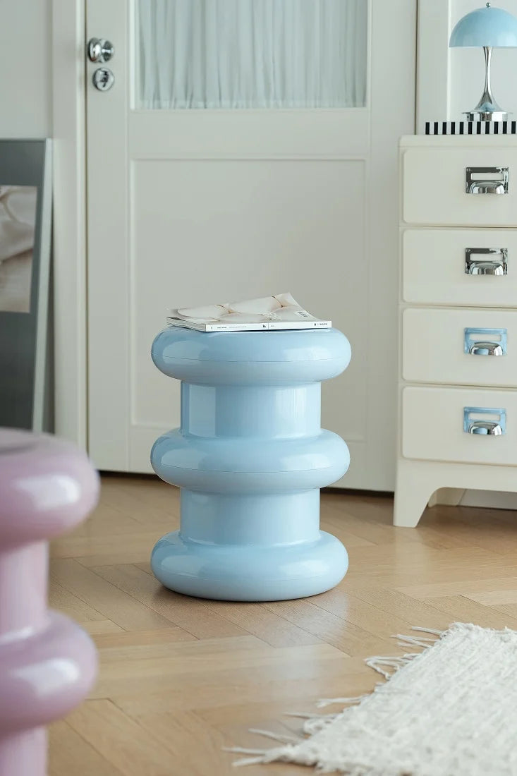 A light blue, tiered barrel-style side table with a glossy finish, styled with a magazine on top in a bright, modern room with light wood floors and white furniture.