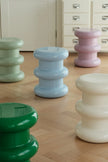 Five glossy, tiered cylindrical stools in pastel green, light blue, lavender pink, cream, and dark green are arranged on a light wood herringbone floor in front of a white cabinet.