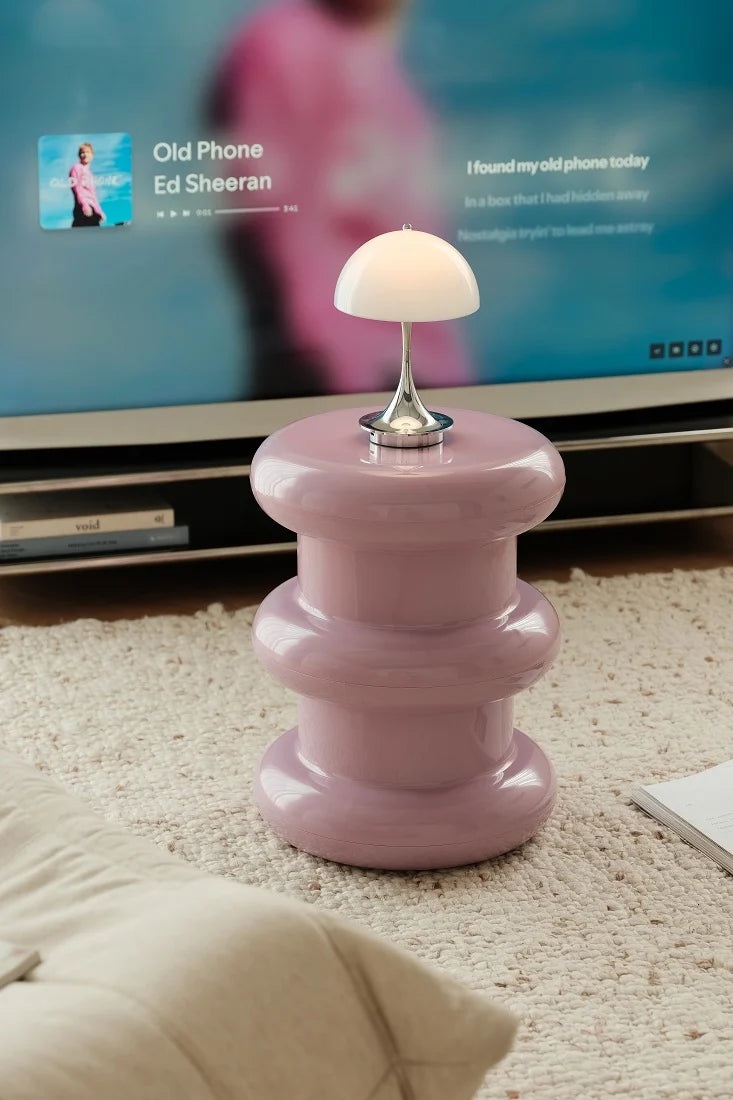 A pink tiered side table lamp with a mushroom-shaped white shade and a silver metal base, set on a beige carpet in front of a television.
