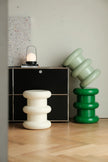 A styled interior scene showing two sculptural, tiered side stools or decor pieces on a light wood floor. One stool is matte white, and the other is glossy green, with a black cabinet and a small lantern in the background against a light wall.