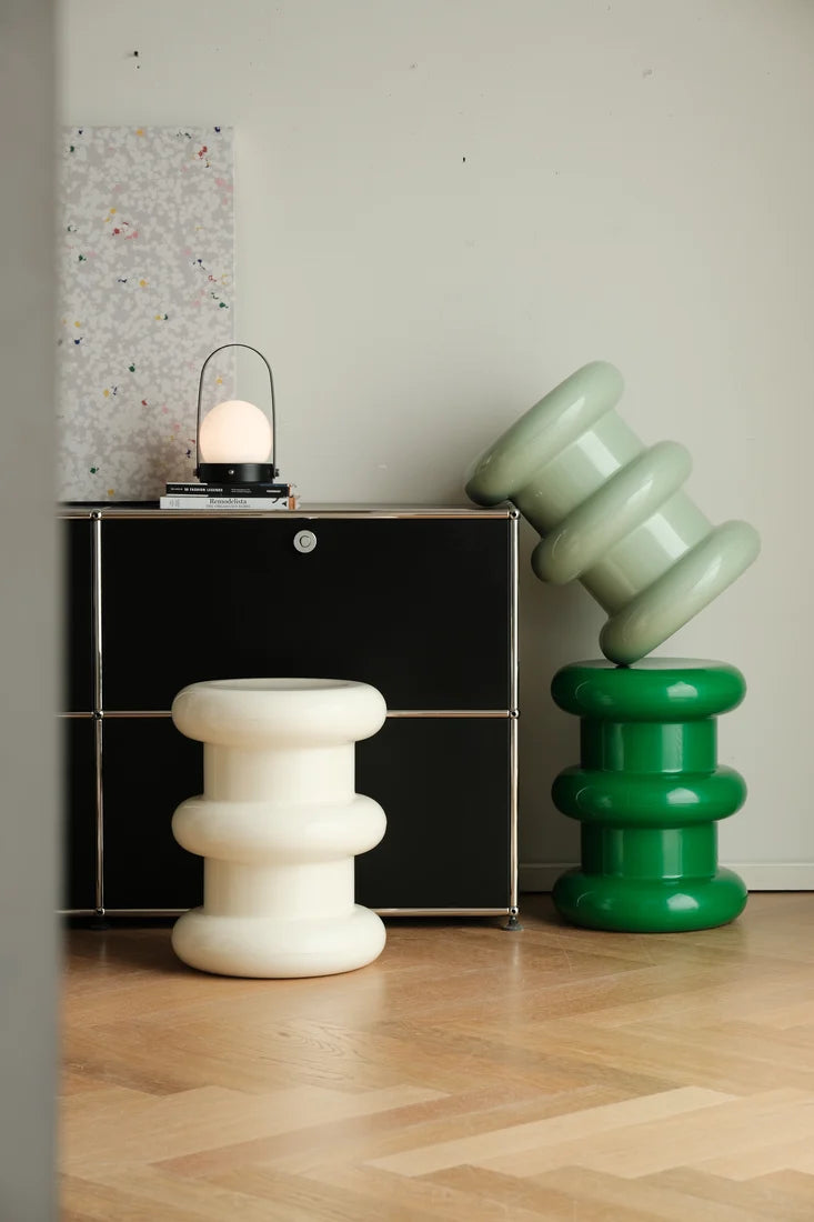 A styled interior scene showing two sculptural, tiered side stools or decor pieces on a light wood floor. One stool is matte white, and the other is glossy green, with a black cabinet and a small lantern in the background against a light wall.