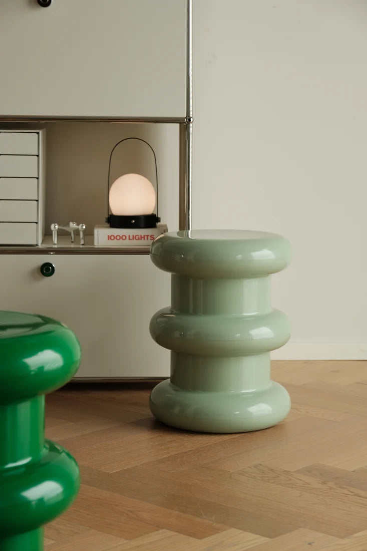 A glossy pale mint green ceramic side stool with a tiered, stacked design sits on a light wood floor next to a white shelving unit with a lit lamp. A portion of another green stool is visible in the foreground.