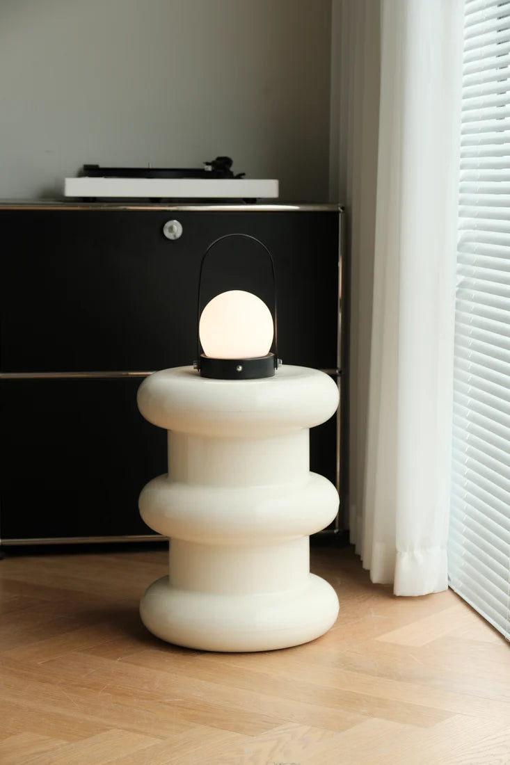 A modern, tiered off-white ceramic side table lamp with a glowing spherical bulb, set on a light wood floor next to a black cabinet and white curtains.