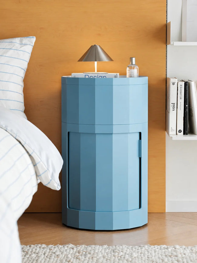A modern, low-profile cylindrical nightstand in a muted light blue color, styled in a bedroom setting next to a bed with white bedding and a wooden headboard.