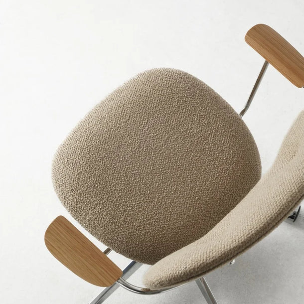 A modern lounge chair with a textured beige bouclé seat and curved backrest, featuring light natural wood armrests and a chrome metal base with casters, photographed on a light gray floor against a white wall.