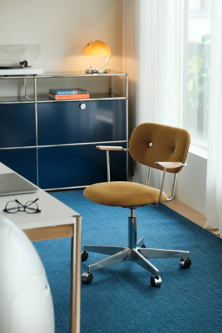 A mid-century modern office chair with a mustard yellow upholstered seat and backrest, mounted on a chrome five-star wheeled base, set in a bright home office next to a blue credenza.