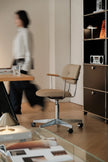 A beige upholstered swivel office chair with a chrome five-star base and armrests, shown in a modern home office setting with a blurred person walking in the background.