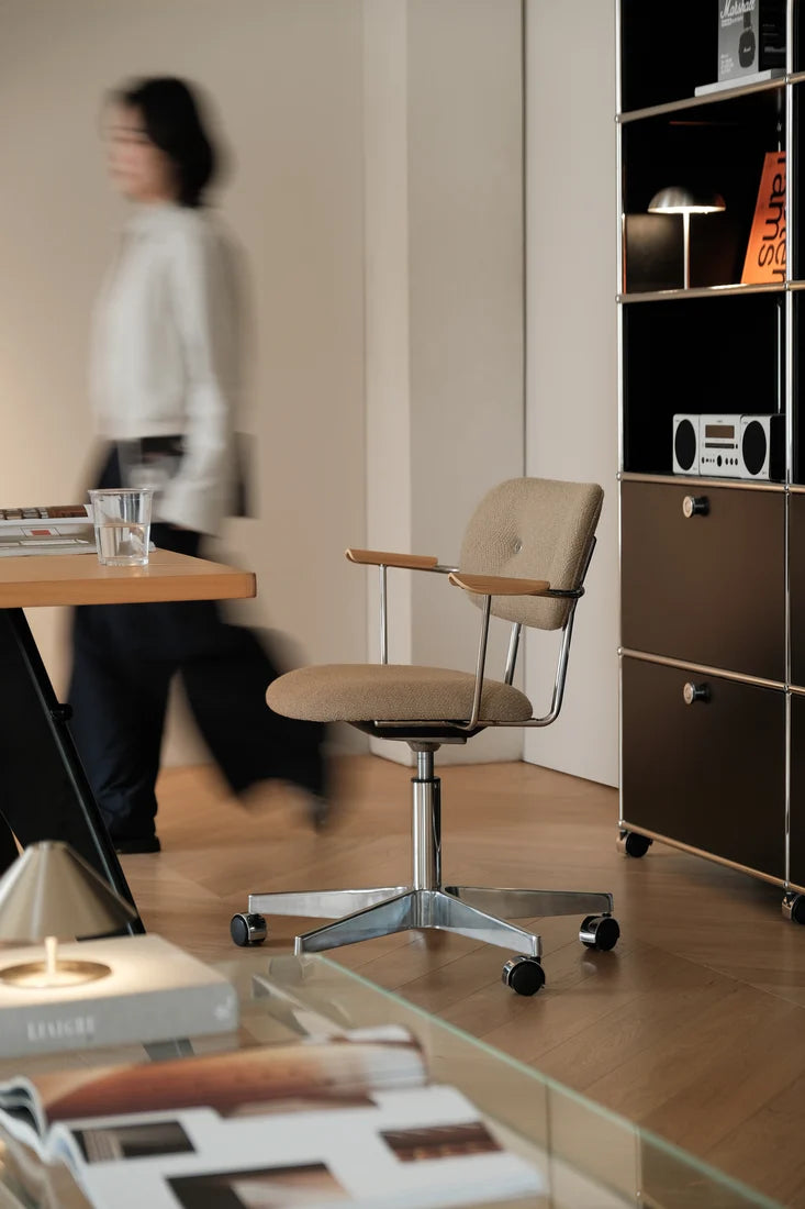 A beige upholstered swivel office chair with a chrome five-star base and armrests, shown in a modern home office setting with a blurred person walking in the background.