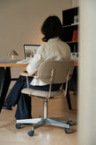 A person sitting on a beige upholstered office chair with a metal frame, viewed from behind in a home office setting.