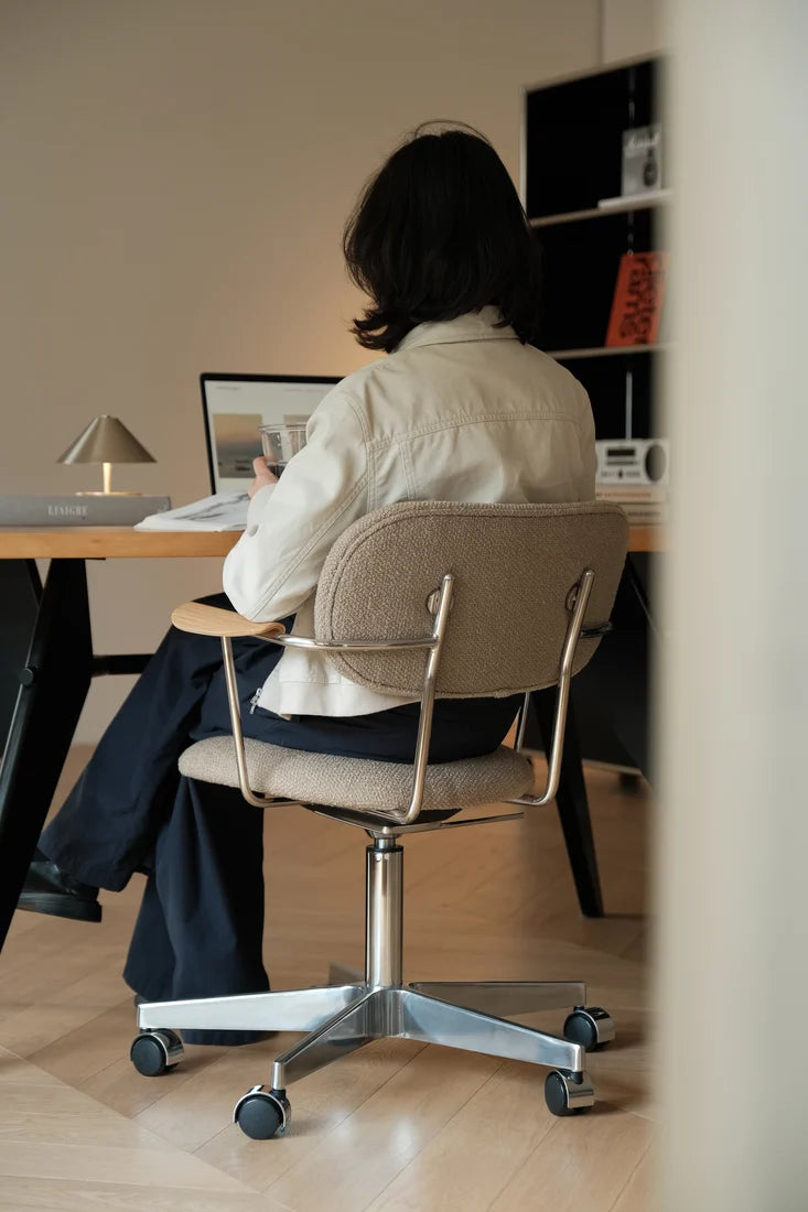 A person sitting on a beige upholstered office chair with a metal frame, viewed from behind in a home office setting.