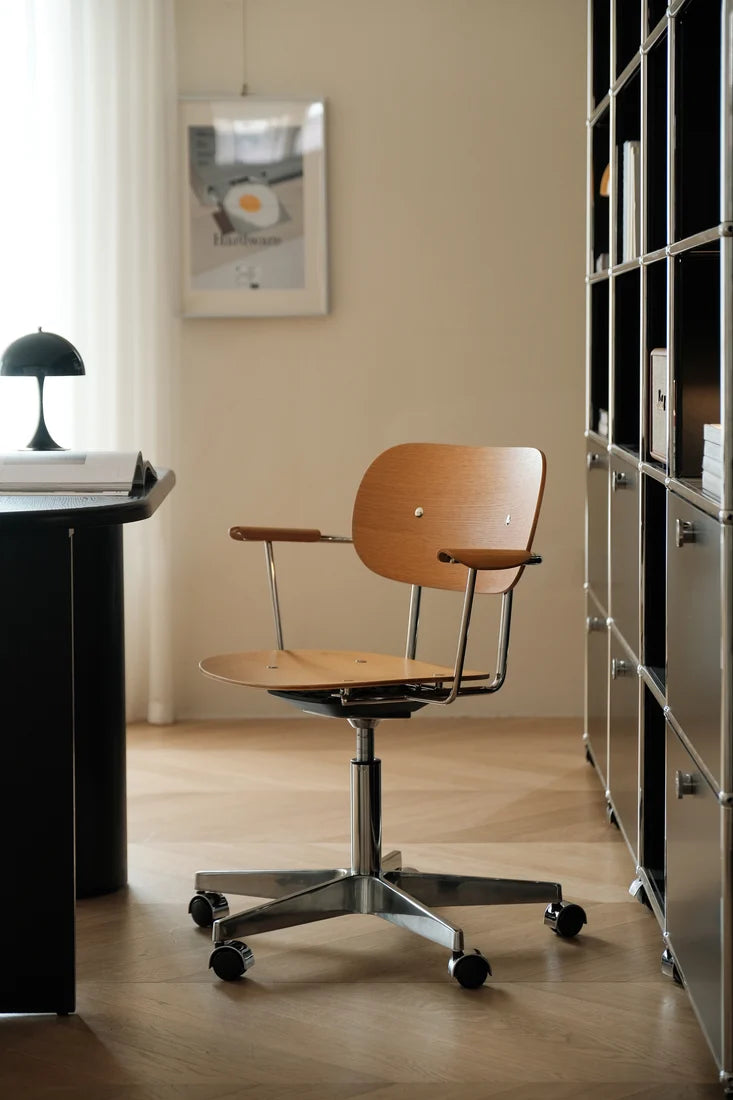 A mid-century modern office chair with a light wood seat and backrest on a chrome five-star wheeled base, positioned in a minimalist home office setting with a desk, shelving, and warm beige walls.