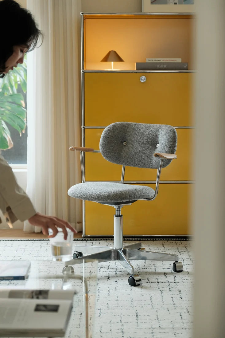 A gray upholstered office chair with light wood armrests and a chrome five-star base, shown in a modern room with a yellow cabinet, window, and a person partially visible on the left.