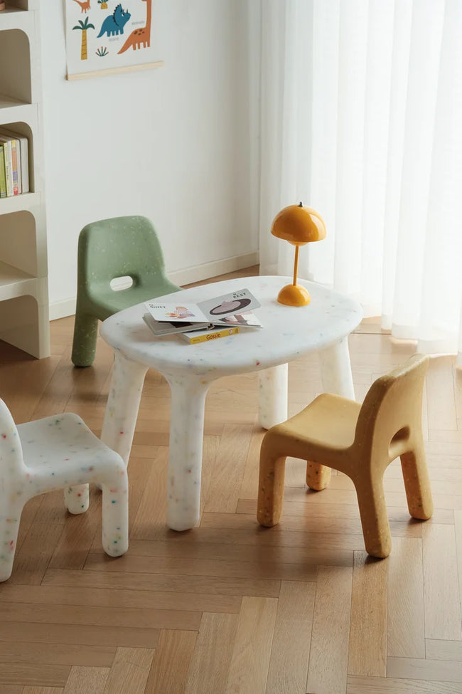 Three small plastic children's chairs in yellow, white, pink, and light blue are arranged on a light wood floor in front of a white plastics desk.