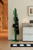 A green cactus-shaped coat rack stands in a modern interior next to a white cabinet, holding a black Chanel shoulder bag and a black scarf with white monogram motifs.