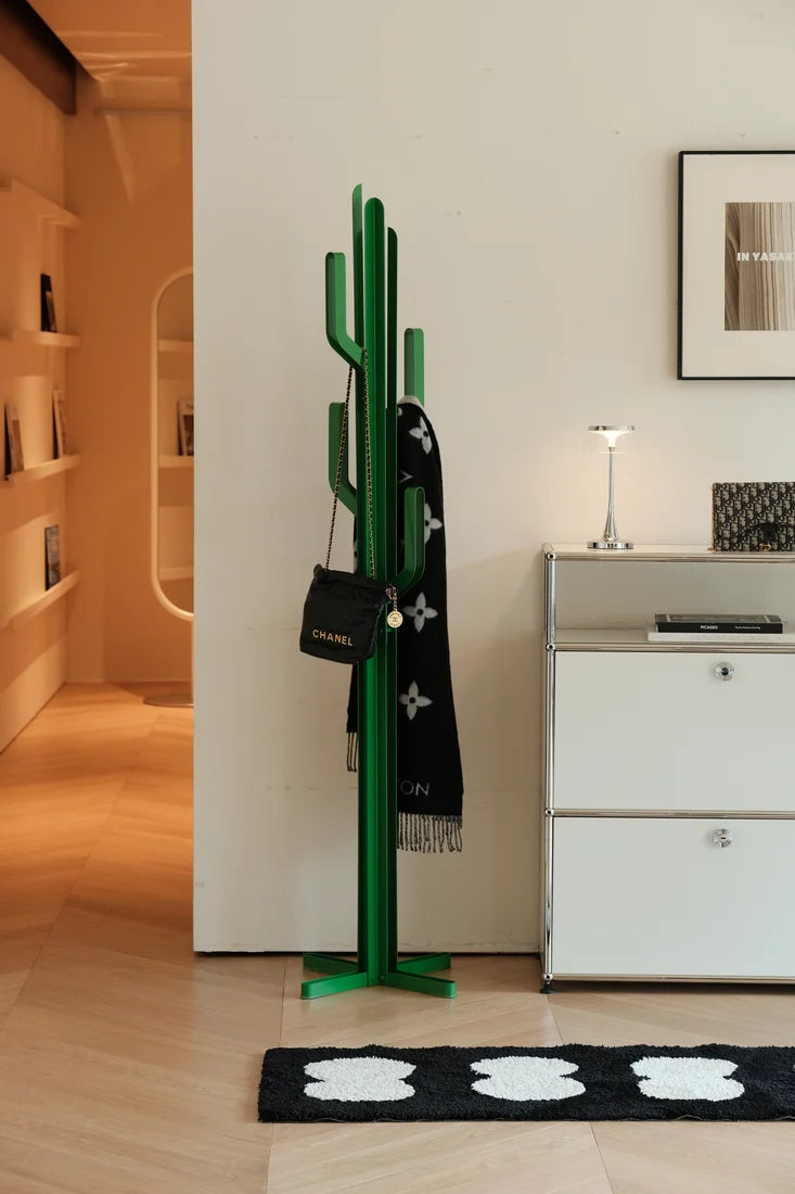 A green cactus-shaped coat rack stands in a modern interior next to a white cabinet, holding a black Chanel shoulder bag and a black scarf with white monogram motifs.