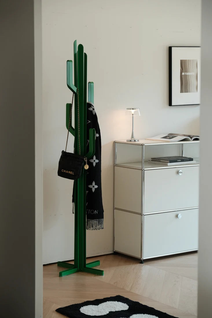 A green cactus-shaped coat rack with translucent green glass hooks stands in a modern room next to a white cabinet, with a black scarf and a black Chanel handbag hanging from it.
