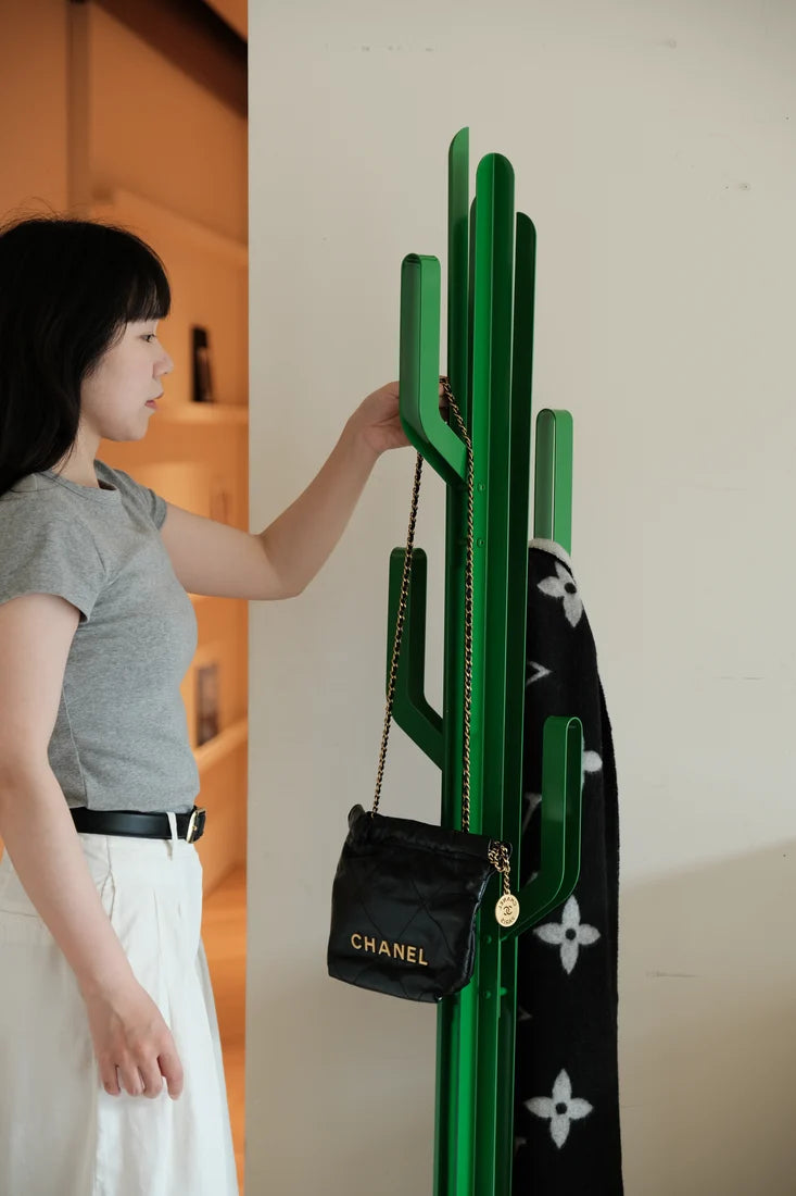 A woman in a gray t-shirt and white skirt stands next to a green cactus-shaped coat rack, holding a black Chanel shoulder bag.