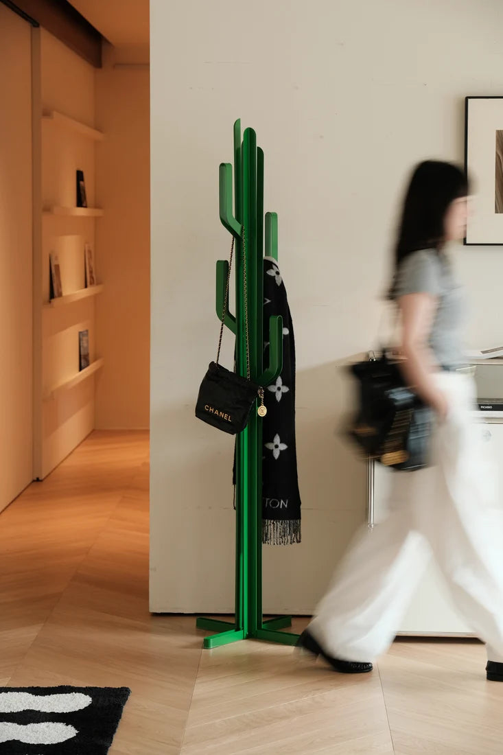 A green cactus-shaped coat rack stands in a modern interior hallway with wood flooring, a blurred person walking past on the right, and a black scarf and handbag hanging from the rack.