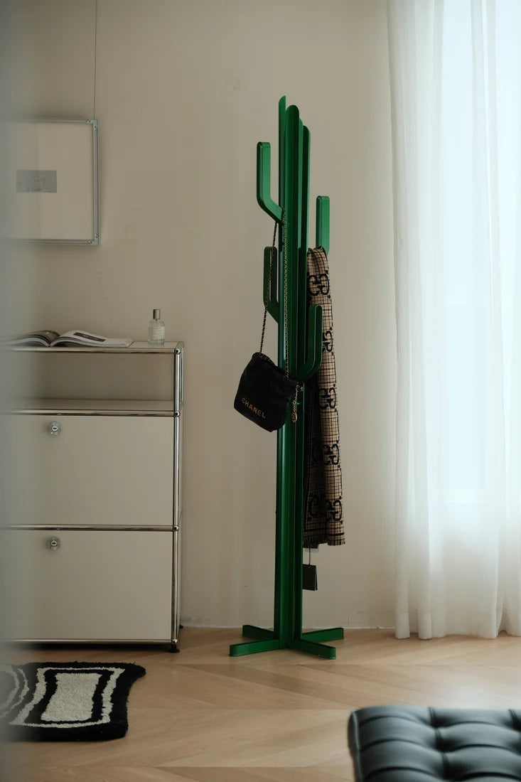 A green cactus-shaped coat rack standing in a bright room with light wood floors, white curtains, and a white cabinet. A black handbag and a patterned coat are hanging from the rack.