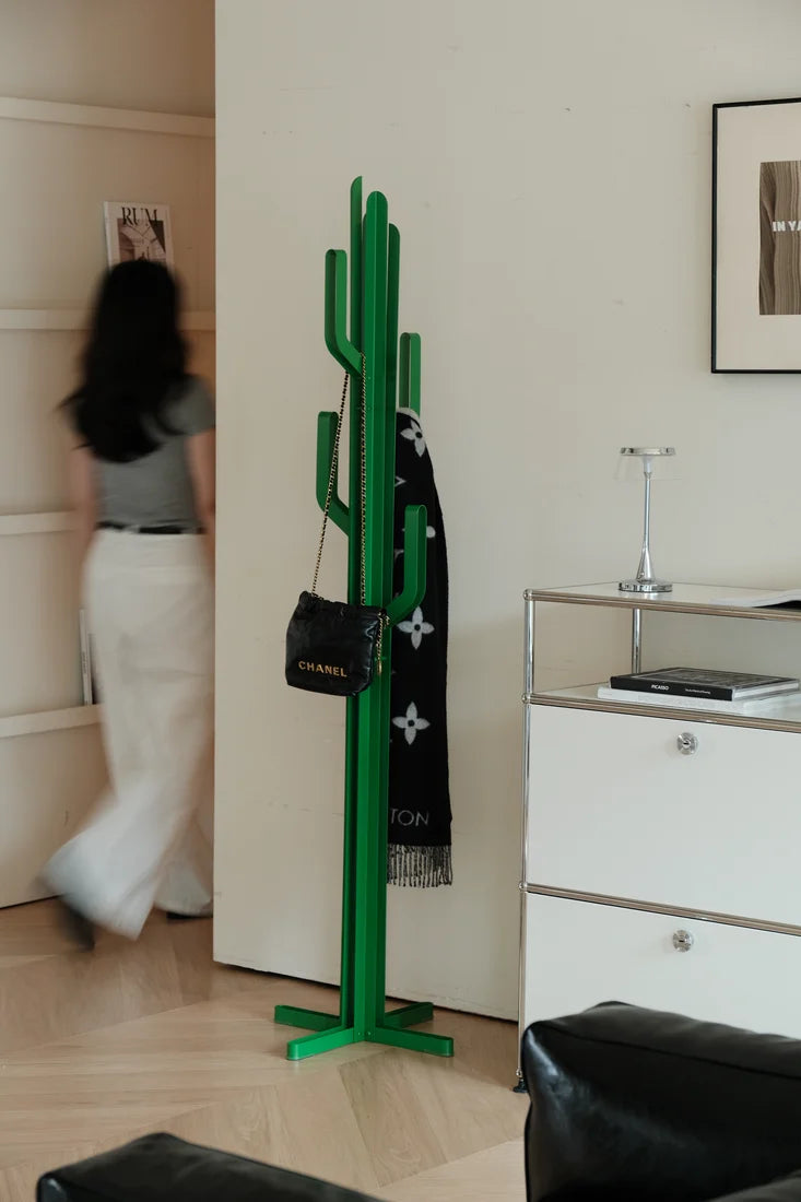 A bright green cactus-shaped coat rack stands in a modern room, holding a black scarf with white monogram-style motifs and a small black handbag with gold lettering. A blurred person walks in the background near a white cabinet.