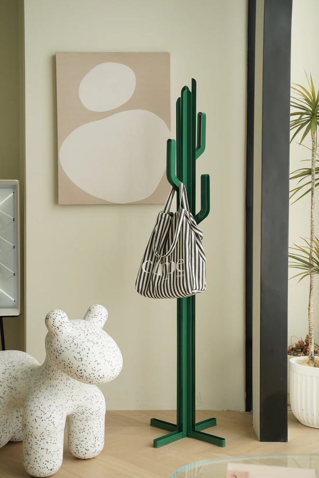 Green cactus-shaped coat rack stand with a striped bag against a light wall with abstract art.