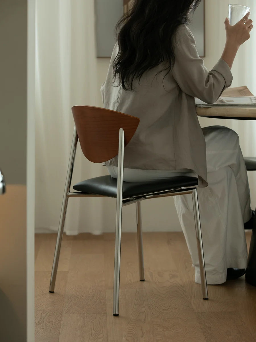 A woman seated at a table in a minimalist interior, viewed from behind. She is wearing a light taupe long-sleeve top and white pants while holding a glass.