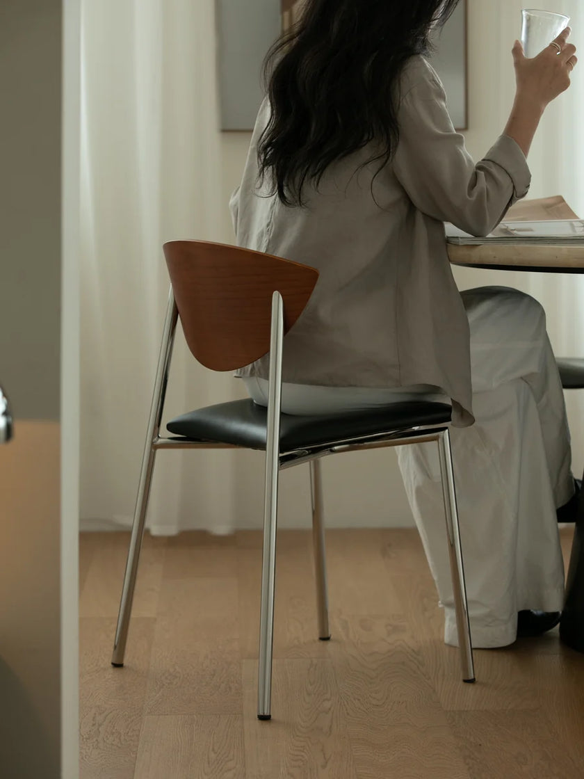 A woman seated at a table in a minimalist interior, viewed from behind. She is wearing a light taupe long-sleeve top and white pants while holding a glass.