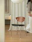 A modern dining chair with a curved wooden backrest, black upholstered seat, and slim metal legs, set in a softly lit room with white curtains and a blurred person in the background.