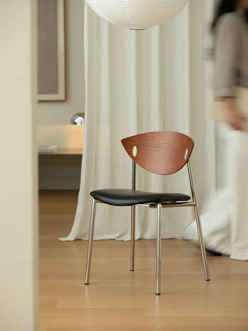 A modern dining chair with a curved wooden backrest, black upholstered seat, and slim metal legs, set in a softly lit room with white curtains and a blurred person in the background.