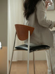 A person seated on a modern chair in a softly lit indoor room, viewed from behind. The stool has a curved medium-brown wooden backrest, a black upholstered seat, and slim silver metal legs.