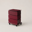 A three-drawer rolling storage cabinet in a deep burgundy finish, featuring a flat top and black caster wheels, photographed against a neutral light beige background.