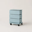 A three-drawer rolling nightstand in a light pastel blue color, featuring a flat top surface and black caster wheels, photographed against a neutral off-white background.