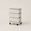 A modern three-drawer nightstand with a white finish, a flat top surface, and black caster wheels, photographed on a seamless light beige background.