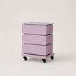 A three-drawer nightstand with a light purple/lavender finish and black caster wheels, shown on a clean, light beige studio background.