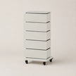 A tall, narrow five-drawer storage cabinet in white with black accents, featuring a flat top and caster wheels at the base, photographed against a plain light beige studio background.