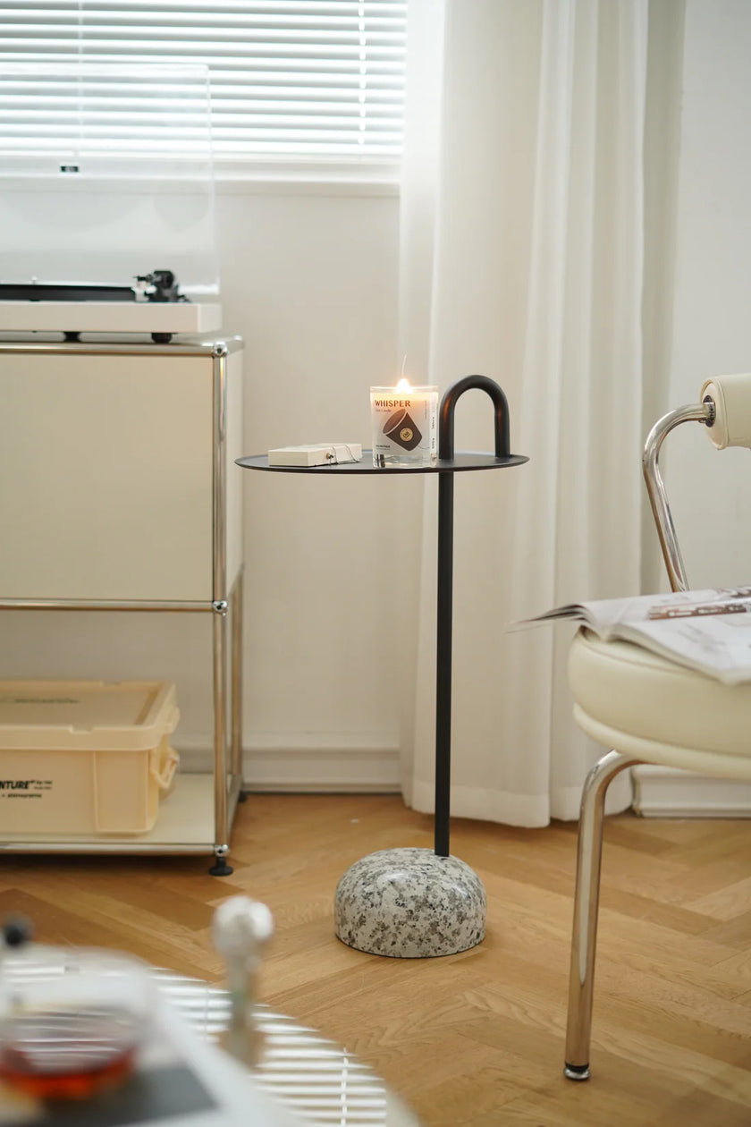 Small round black side table with a black metal frame on a wooden floor next to a chair and a window with blinds.