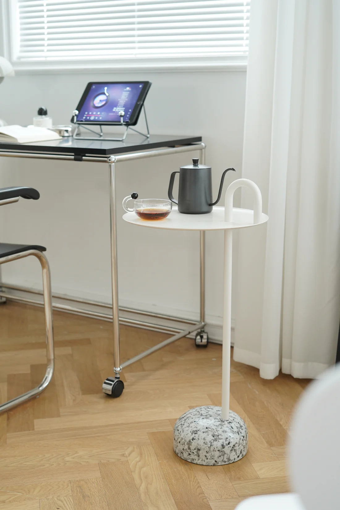 Small white round side table with a teapot and cup on a wooden floor next to a desk.