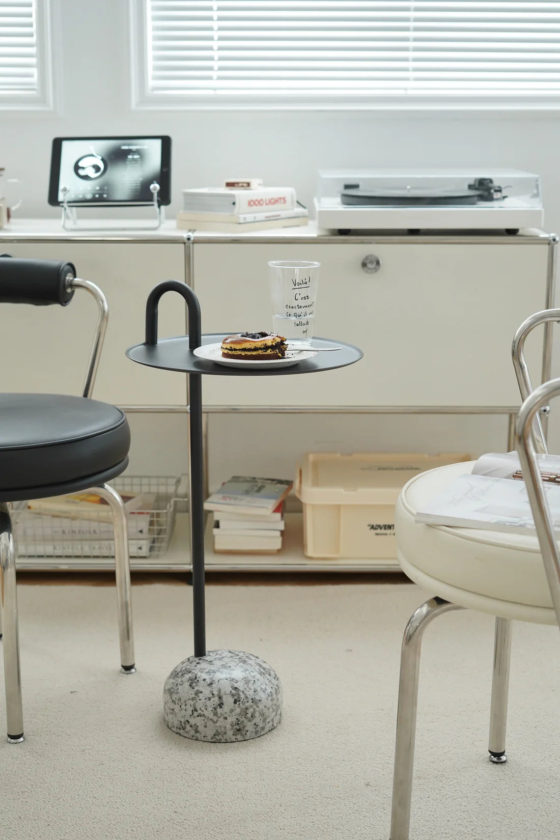 Small Black round side table with a glass of water and a donut on a shelf in a room with chairs and a computer.