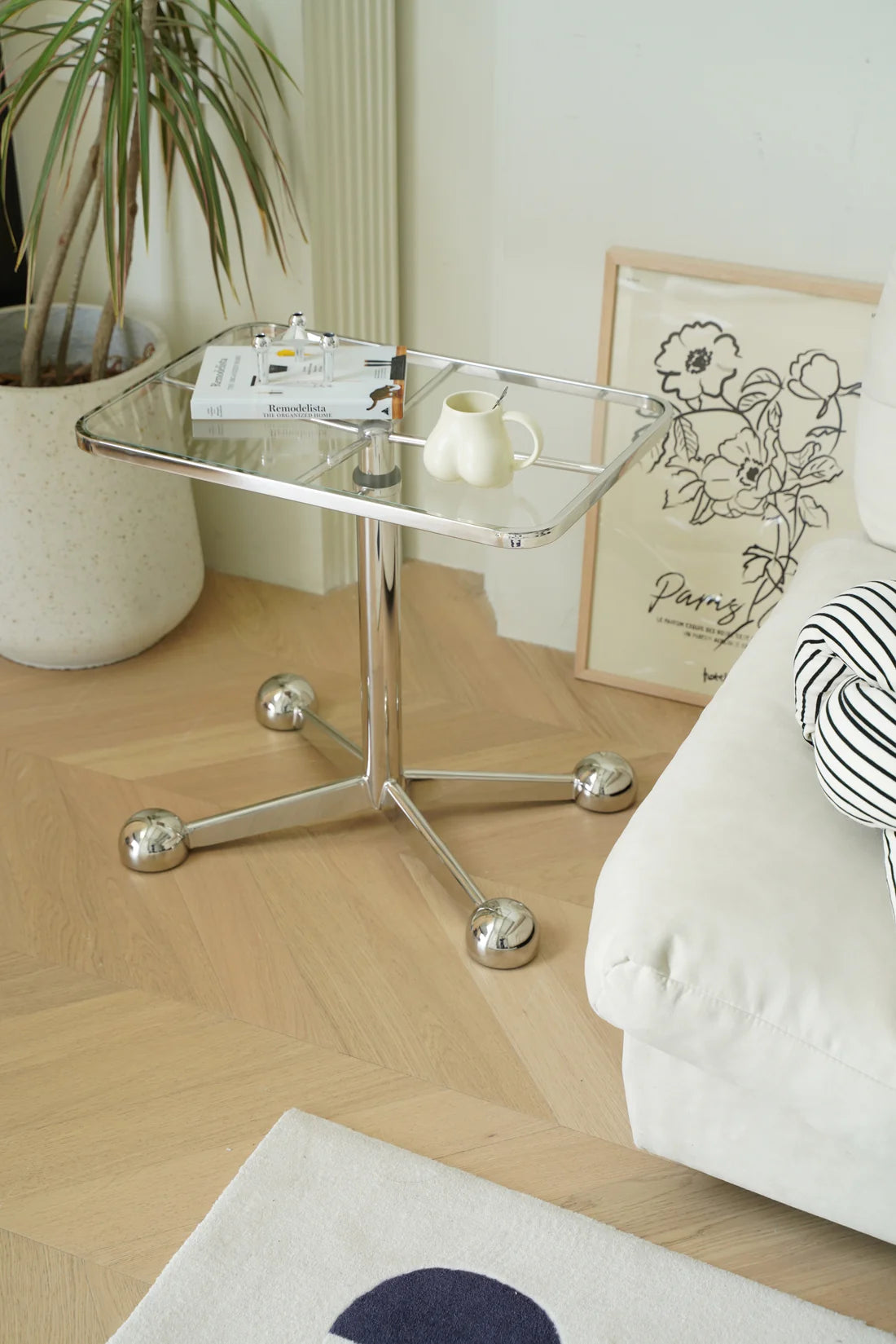 Glass sofa side table with a plant, books, and a cup in a living room setting.