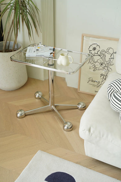 Glass sofa side table with a plant, books, and a cup in a living room setting.