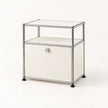 A modern modular storage cabinet with a chrome metal frame, a clear glass top shelf, a cream-colored middle shelf, and a cream lower door with a small round lock. The cabinet stands on black feet against a clean, off-white background.
