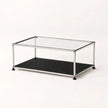 A modern two-tier coffee table with a clear glass top, a black lower shelf, and a slim stainless metal frame with black feet, photographed on a seamless white background.